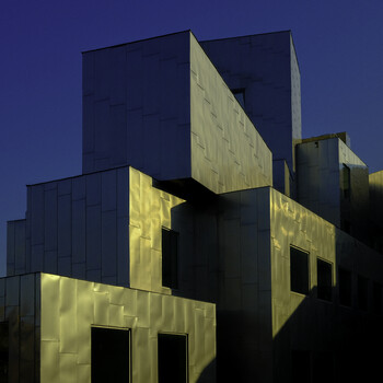 Modern angular building with yellow-lit facades and dark exterior walls against twilight sky.