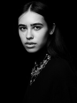Black and white portrait of a young woman with long dark hair and direct gaze against dark background.