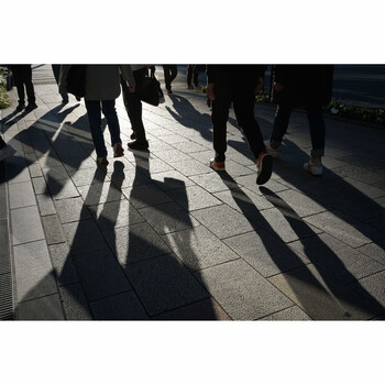 Silhouettes of pedestrians cast across a paved street in bright sunlight, creating dramatic shadows.
