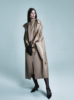 Woman in neutral beige trench coat against plain blue-gray backdrop.
