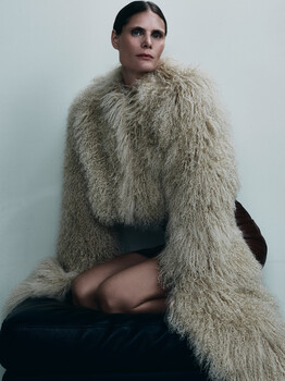 Woman in knit sweater and oversized fur stole seated against neutral background.