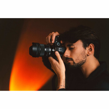 Male photographer holding a camera to his face in profile against warm orange and dark background lighting.