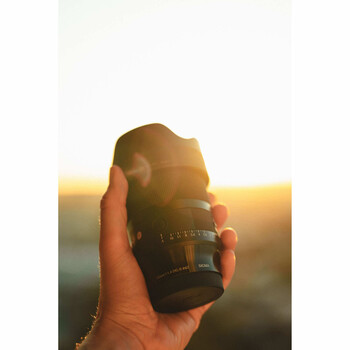 Hand holding a telephoto lens backlit by golden sunset light with blurred landscape behind.
