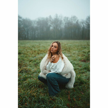 Woman in cream cardigan crouching in misty field with coniferous forest background.
