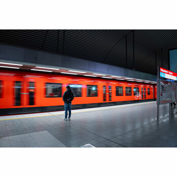 A lone commuter stands on an empty subway platform beside a vibrant red-orange train.