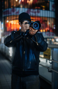 Photographer holding a DSLR camera up to eye level on an urban street at night.
