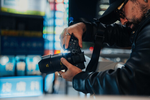 Photographer adjusting settings on a mirrorless camera with urban buildings blurred in background.