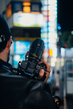 Photographer holding a telephoto lens while shooting at night in an urban environment with blurred city lights in the background.