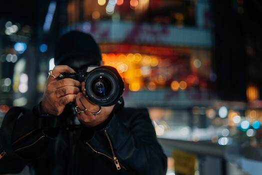 Photographer holding a DSLR camera with lens extended, shooting at night with city lights blurred in background.