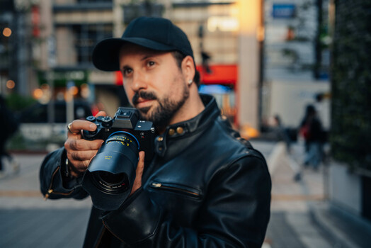 Photographer in black jacket and cap pointing camera toward viewer on urban street.