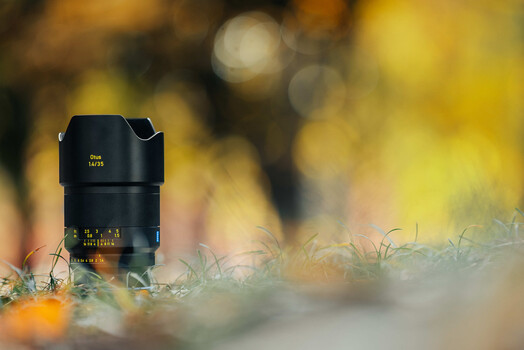 Black camera lens with manual focus ring mounted on mossy surface with warm, blurred bokeh background.