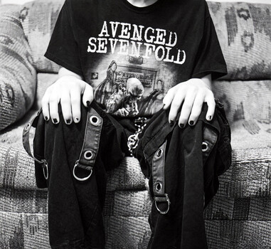 Person wearing Avenged Sevenfold band shirt and studded black accessories posed on patterned couch.