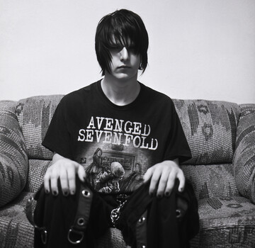 Person in black band t-shirt and studded accessories seated on gray couch in black and white portrait.
