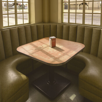 Minimalist interior photograph of a rounded rectangular table with light wood top and dark pedestal base in a booth with olive-green upholstered walls and large gridded windows.