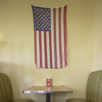 American flag tapestry hanging on a pale yellow wall above a small round table with two green chairs.