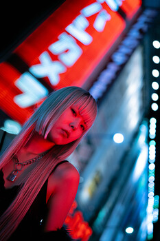 Woman with long hair lit by neon red and blue light against blurred signage backdrop.