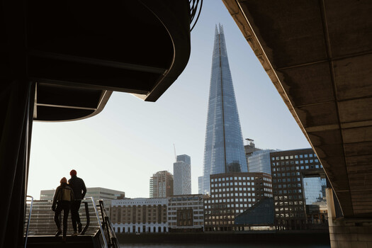 Two figures silhouetted in a concrete structure frame looking toward a tall glass skyscraper amid urban architecture.