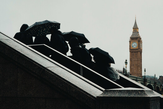 Silhouetted figures with umbrellas walking down a wet roof, with a clock tower visible in the background.