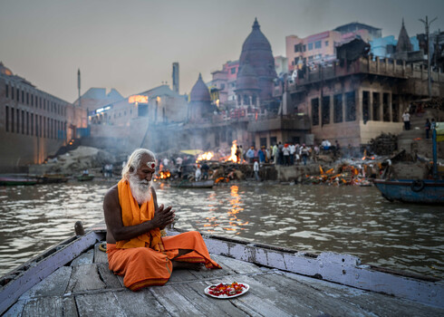 Elderly man in orange robes praying on a boat at dawn, with riverfront temples and ghats visible across the water.