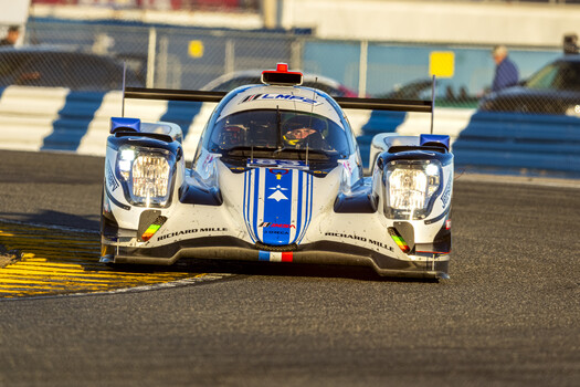 White and blue racing prototype car with dual headlights navigates a banked turn on a race track.
