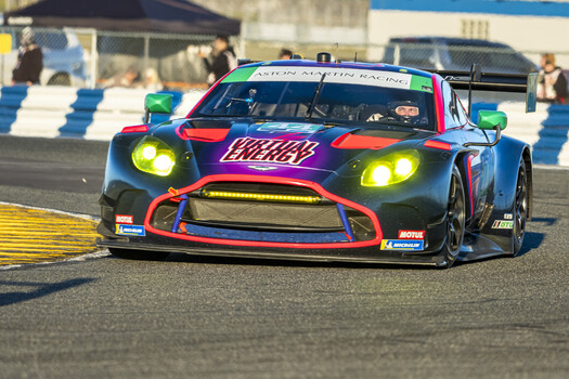 Race car with vibrant purple and blue livery accelerates on a circuit track with yellow curbing.