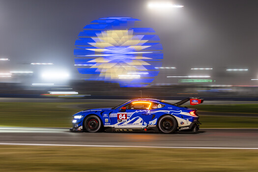 Blue race car number 84 photographed with motion blur on a nighttime track with illuminated signage.