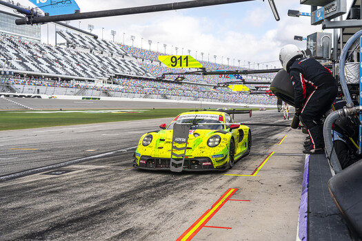 A bright yellow race car number 911 positioned on pit lane at a major motorsport facility with grandstands visible in the background.