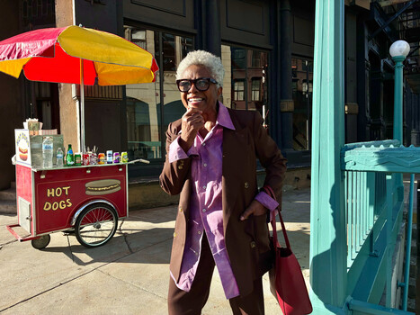 Elderly woman in purple jacket and glasses posing on sunny street near hot dog cart.