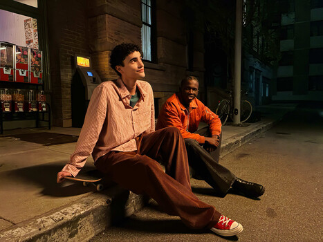 Two people sitting on a curb at night in an urban alley, lit by warm artificial light from storefronts.