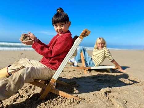 Child in red sweater using wooden shovel to dig in sand on beach with another child visible in background.