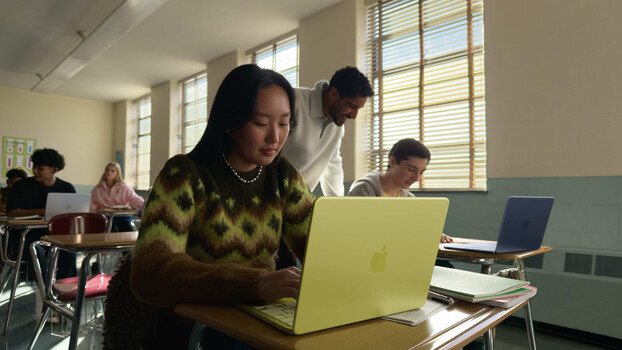 Woman in patterned sweater working on yellow laptop at desk in bright classroom with other students.