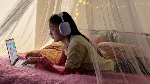 Person wearing wireless headphones lying on a pink rug under sheer draped fabric, working on a laptop.