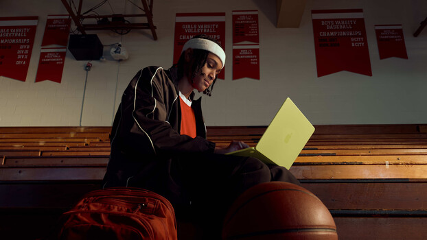 Person sitting on basketball in gymnasium holding yellow notepad, wearing white headband and dark jacket.