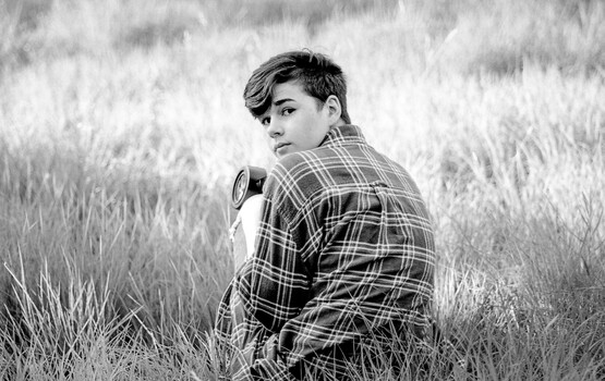 Young person in plaid shirt holding camera, photographed in profile against tall grass field.