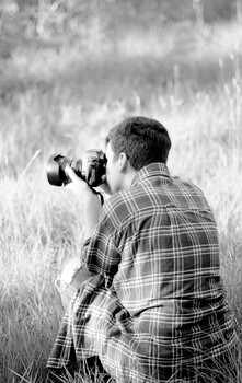 Photographer crouched in tall grass, holding camera to face while wearing plaid shirt.