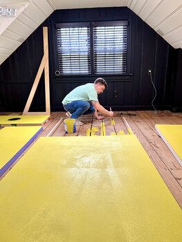 Worker installing bright yellow insulation material on wooden floor joists in an attic space.