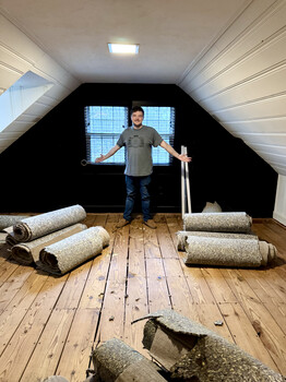 Man standing in attic space with rolled insulation materials on wooden floor.