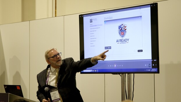 Man in business casual attire pointing at a large display screen showing a presentation with a logo during what appears to be a conference or speaking event.