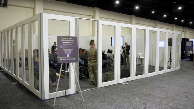 Glass-walled exhibition booth with multiple visitors and a purple informational sign at the entrance.