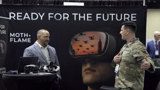 Military personnel in camouflage uniform speaking with civilian at exhibition booth displaying virtual reality headset.
