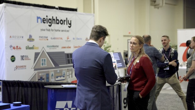 Two professionals conversing at a home services trade show booth with sponsor logos displayed.