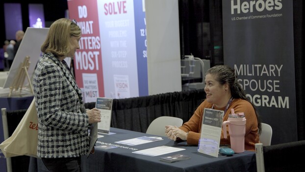 Woman standing at a conference booth speaking with a seated attendant at a table displaying materials about a military spouse program.