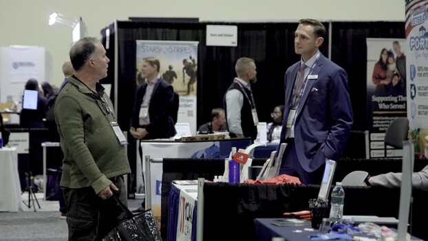 Two professionals conversing at a trade show booth with exhibition banners and displays visible in the background.