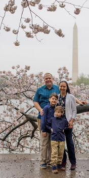 Family of four posing outdoors beneath cherry blossom branches with Washington Monument visible in background.