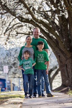 Family of four wearing matching green shirts standing together on a tree-lined path.