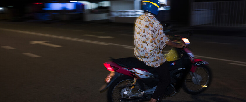Motorcyclist riding at night with motion blur, wearing patterned shirt and cap.