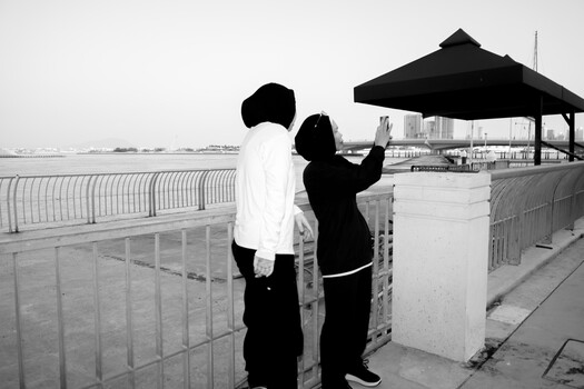 Two people standing at a waterfront railing, one pointing toward the distant cityscape under an overcast sky.