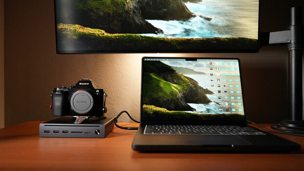 Photographer's desk setup with Sony camera, USB hub, laptop displaying landscape wallpaper, and dual monitors.