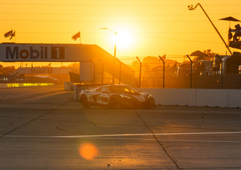 Racing car silhouetted against golden sunrise on a circuit track with barriers and spectator structures.