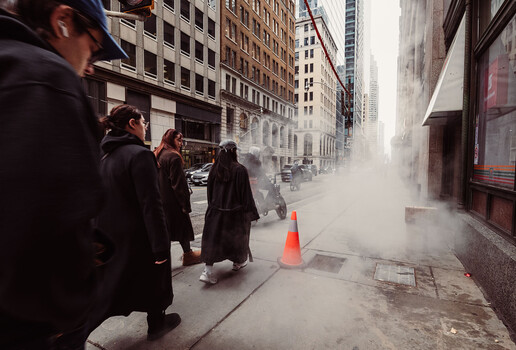 Urban street scene with steam venting from underground grate as pedestrians walk past tall buildings.